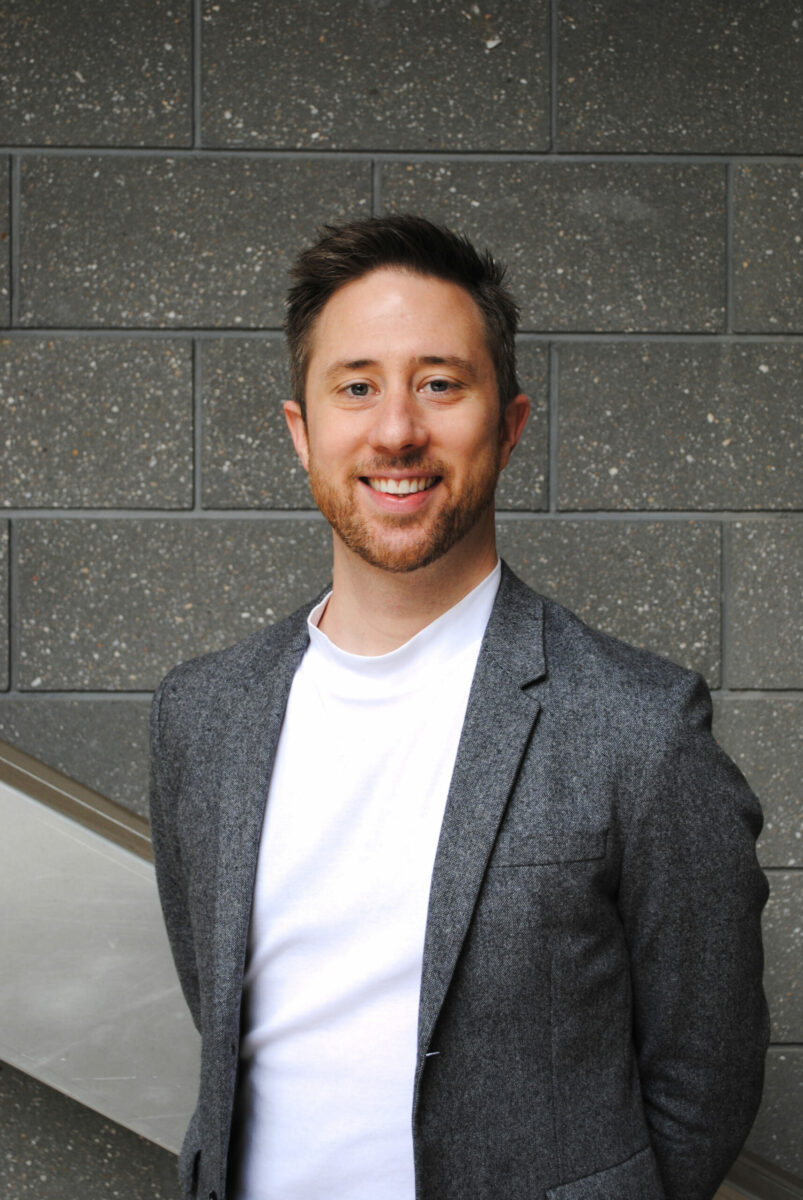 A man smiling wearing a t-shirt and blazer.