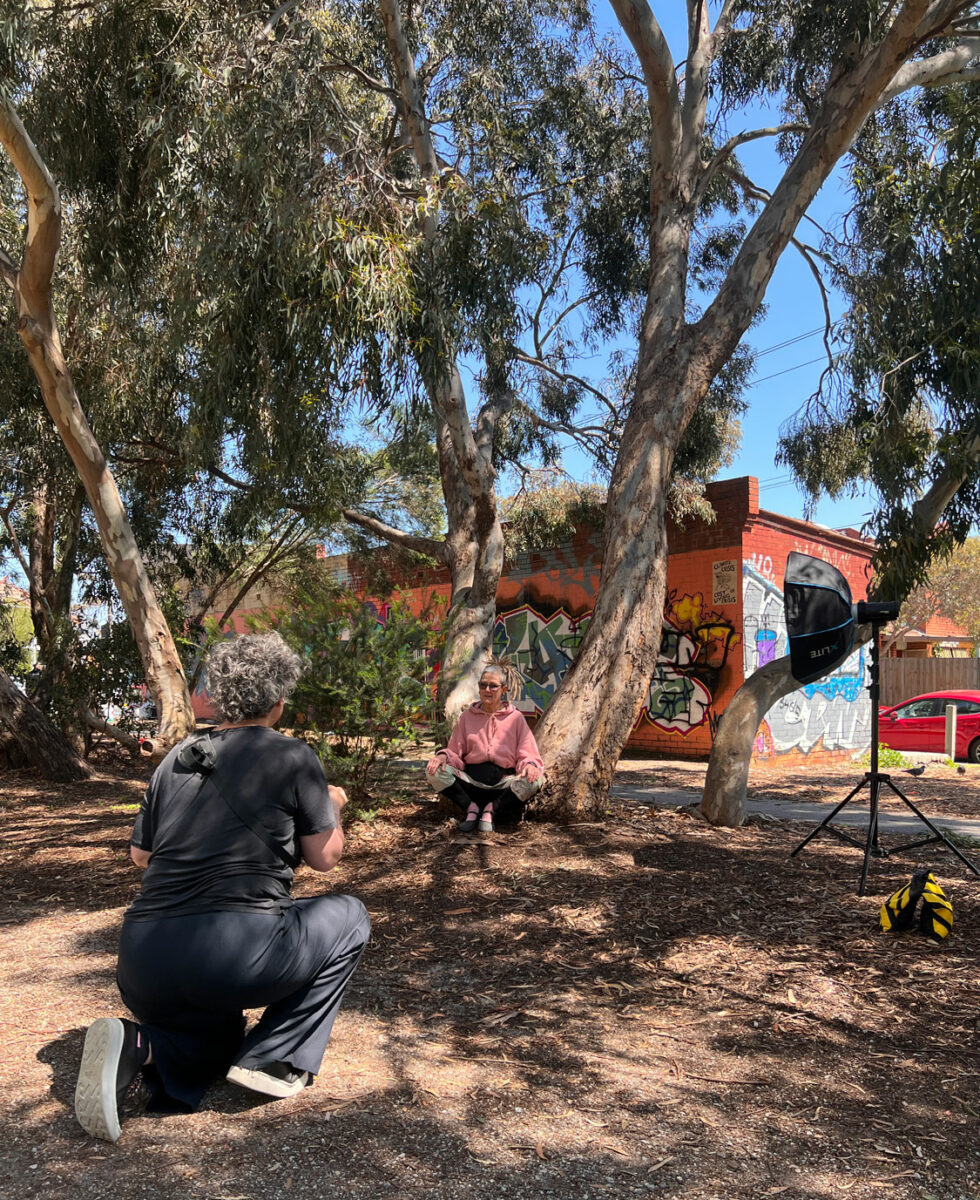 A woman prepares to take a photo of another woman who is seated by a tree..