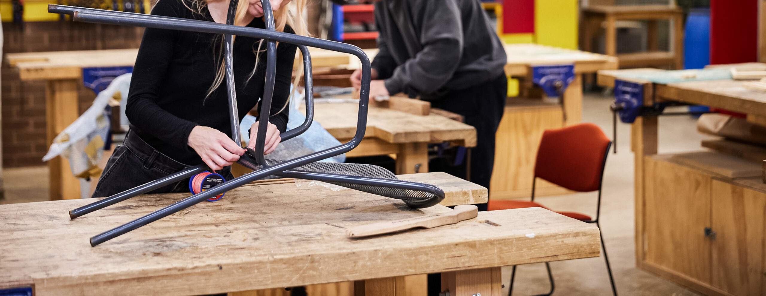 Woman in foreground mending chair on table. Man in background with headphones on.